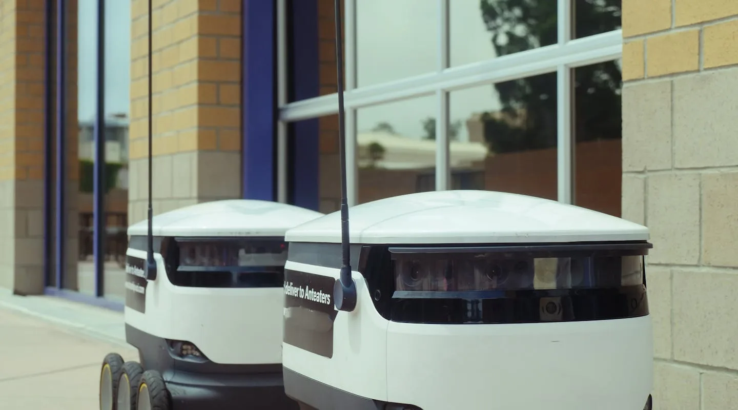 Autonomous delivery robots parked beside a building, equipped with cameras and sensors for sidewalk navigation