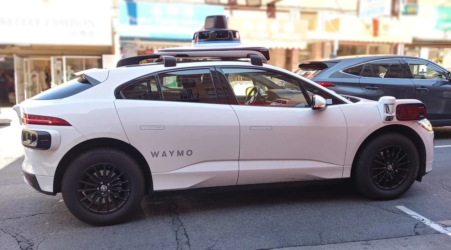 A Waymo self-driving car with visible LiDAR sensor on the roof, driving on a city street
